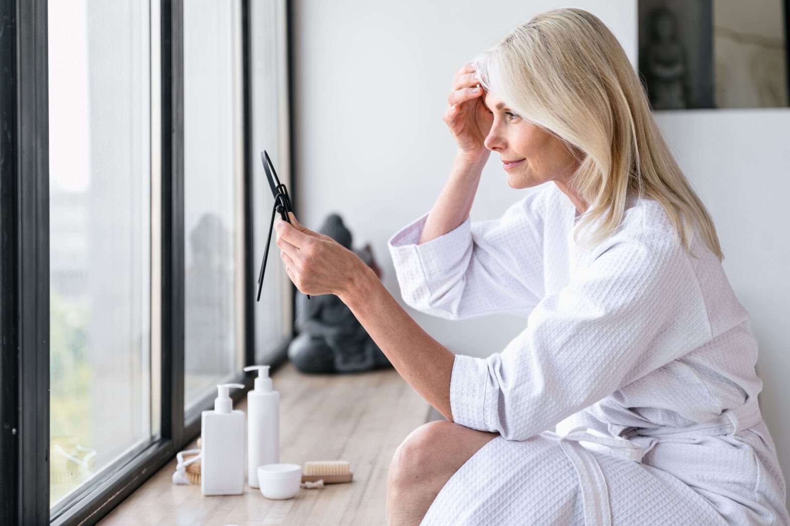 middle aged woman in white robe touching face while looking into mirror post facial plastic surgery skincare washington dc